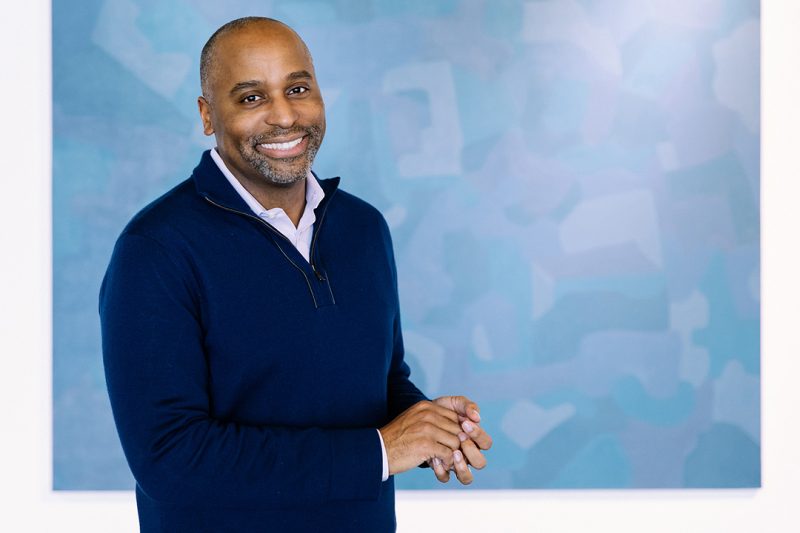 Headshot for Harvey Young, a black man in a sweater smiling and standing in front of a blue background