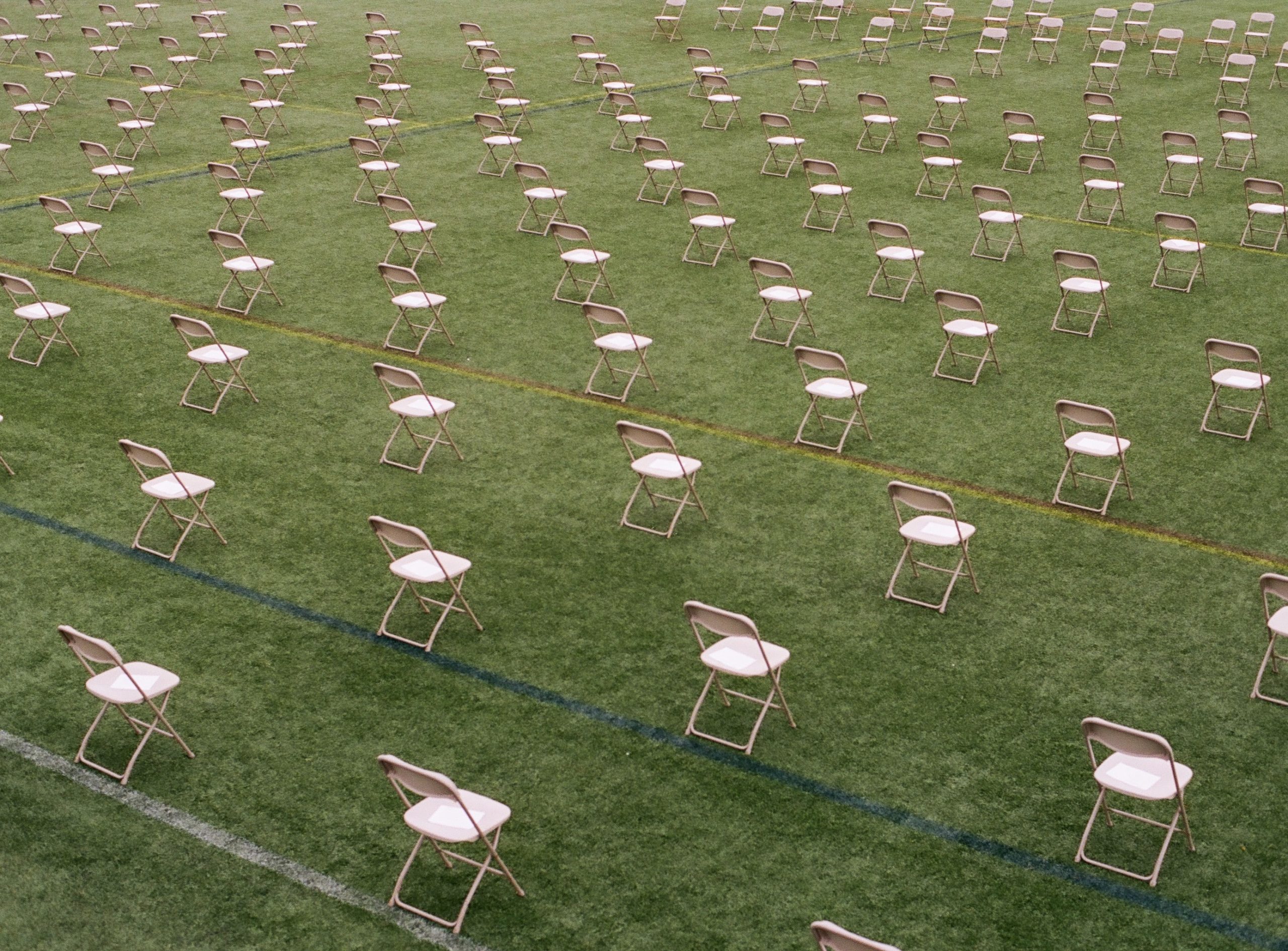 A photo looking down at several rows of pink picnic chairs placed six feet apart and facing away from the camera. The chairs are in an outdoor sports field with white, blue, yellow, and red lines painted on the grass.