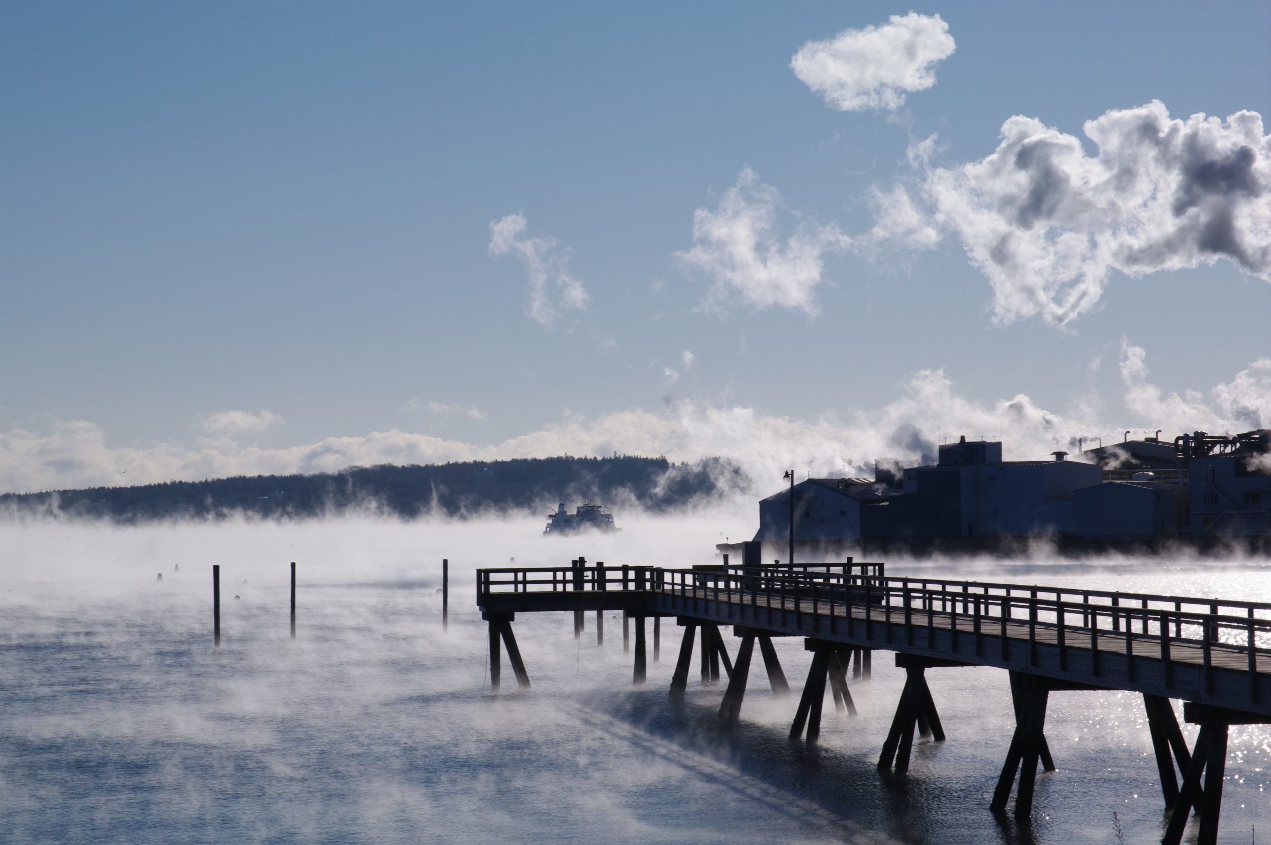A photograph of a long wooden dock on a frozen lake. On the far right, a large factory is depicted. The lake is covered in frost smoke and the sky is a light blue with whisps of white clouds.