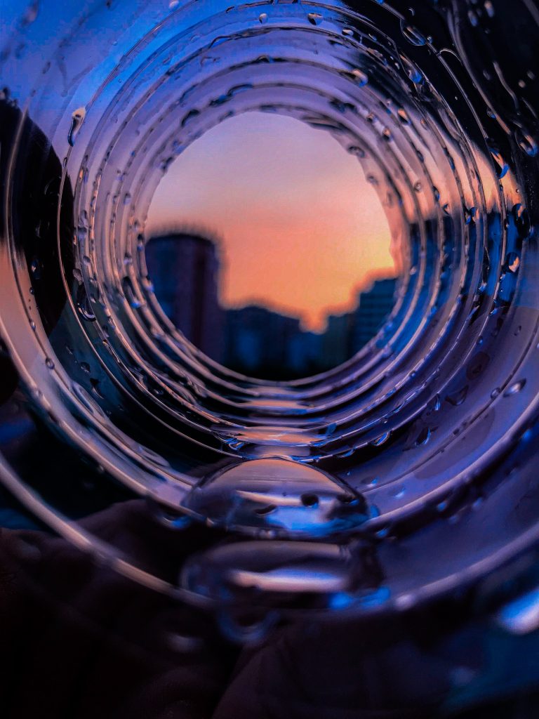 A photograph taken from the inside of a wave of water. The wave forms a momentary translucent tunnel of water. At the end of the water tunnel, the blurry outline of a skyscraper against an orange sunset is depicted.