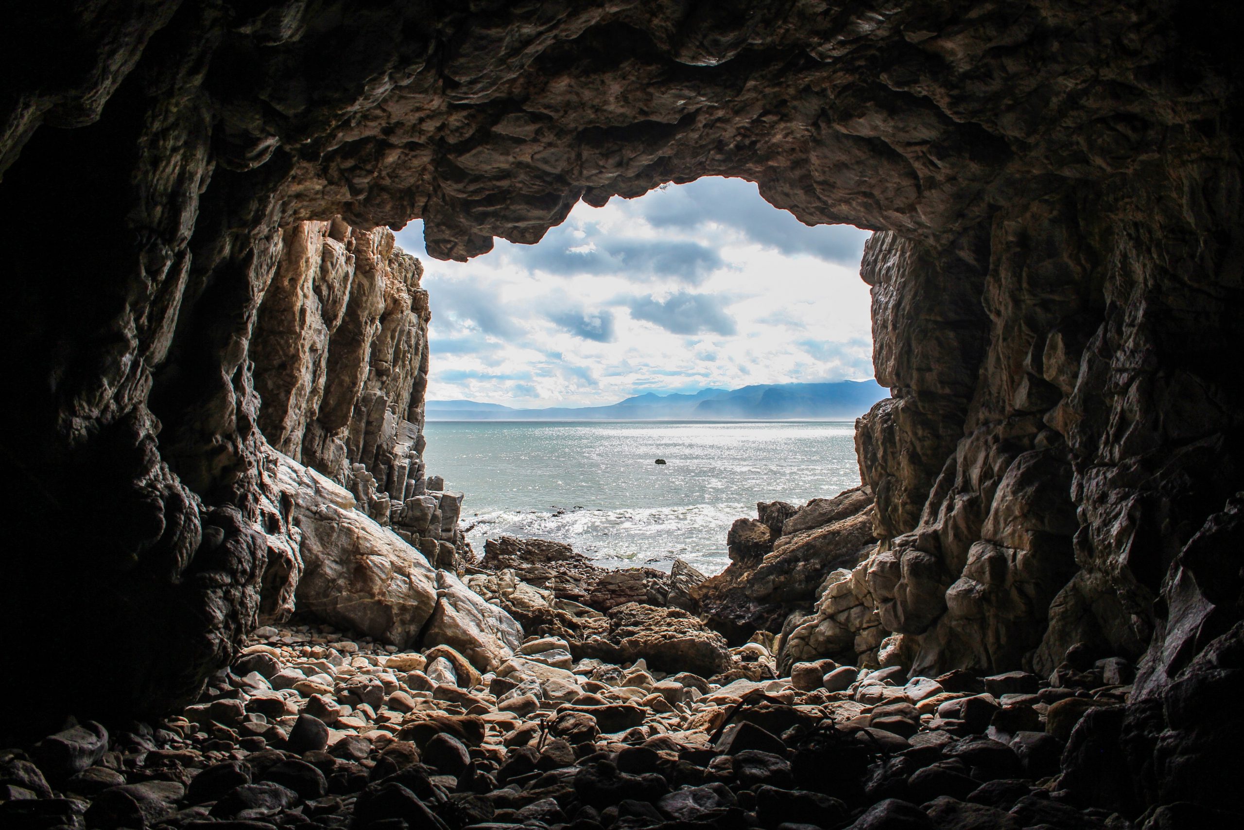 A photograph of the opening of a rocky cave. The camera points outwards from within the cave, depicting a shimmering blue lake, grey clouds, and mountains in the distance.