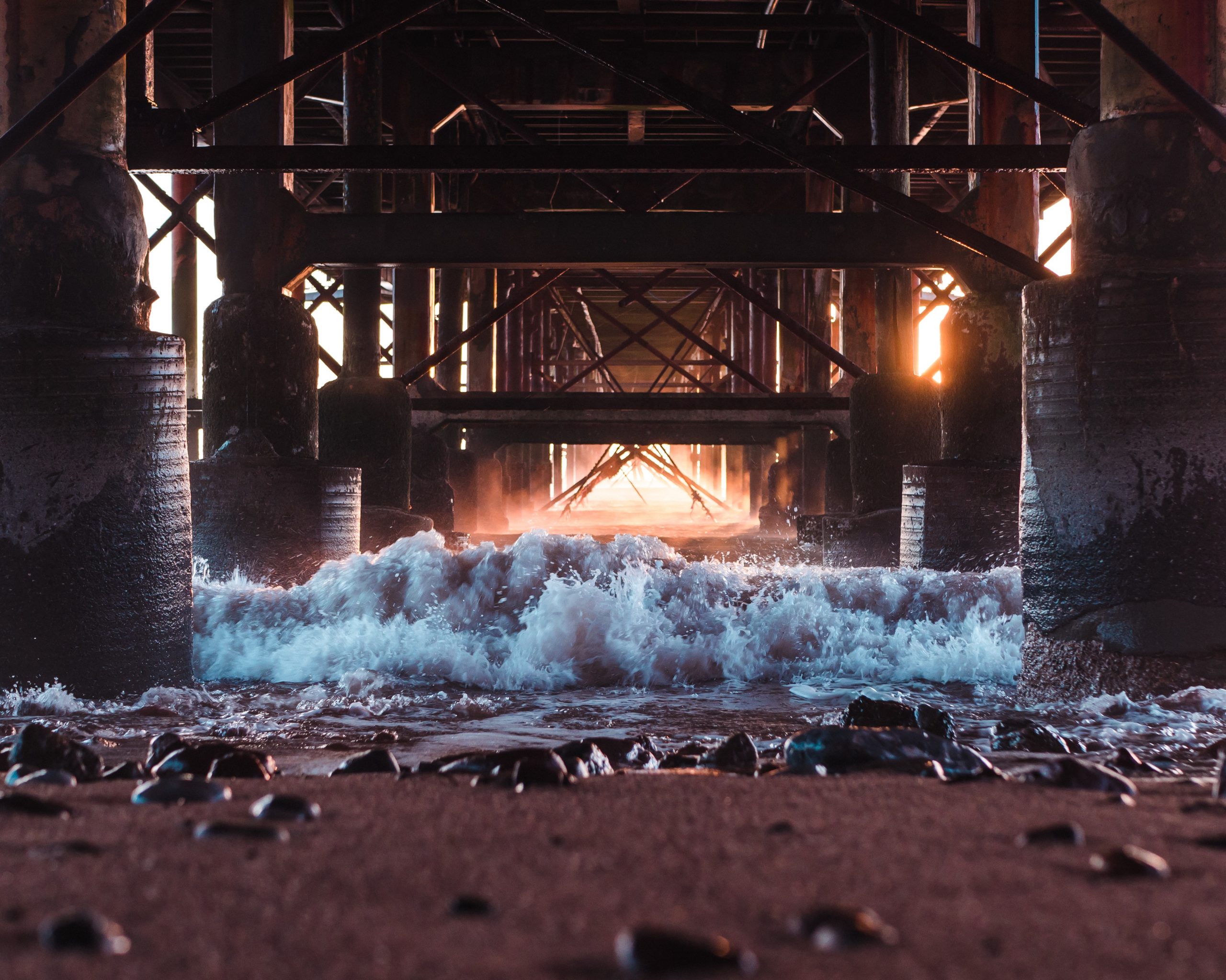 A photograph taken beneath a large boardwalk. Part of a sandy beach with black stones, and an incoming crashing wave is depicted in the front part of the photo. In the distance, the rising sun shines through the gaps in the boardwalk.
