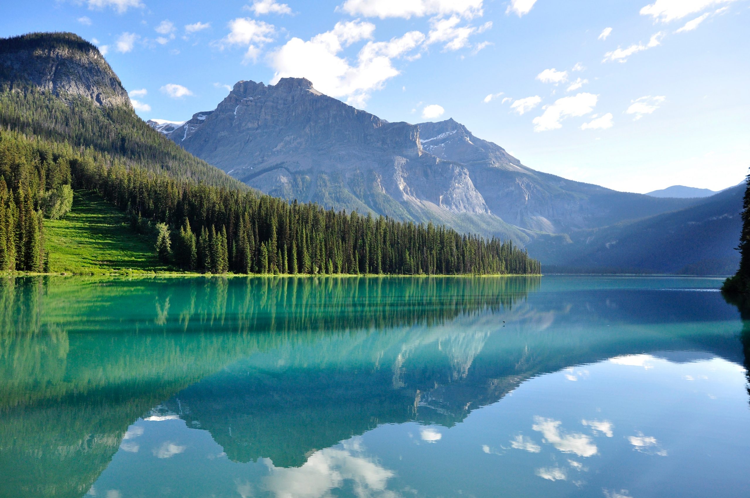 A photograph of a serene, still lake in front of a jagged rocky mountain. On the left, the rocky cliff face is covered by a dense, green pine forest. The water in the lake is so still it clearly reflects the jagged mountain, the blue sky, and the wisps of white clouds in the sky.