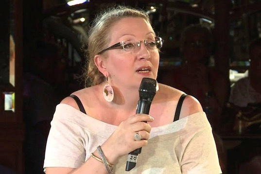 Une photo de tête d'une femme blanche aux cheveux blonds, avec des lunettes fines et un chandail blanc. La femme tient un microphone noir près de sa bouche et est photographiée en train de parler dans ce microphone.