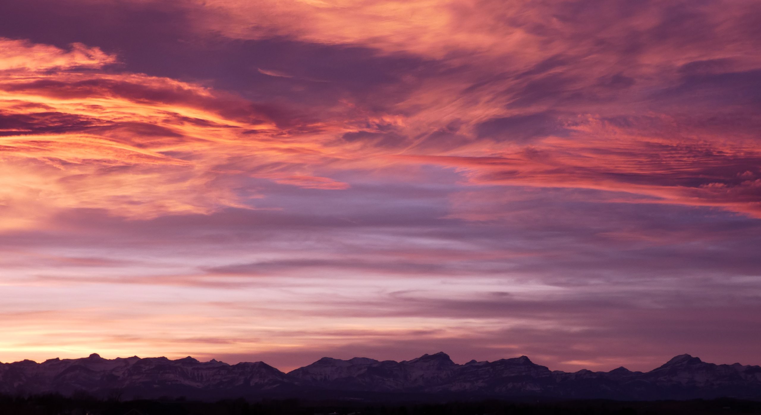 Une photographie d'un coucher de soleil calme. Au bas de l'image, on voit une longue ligne de sommets de montagnes déchiquetés. Au-dessus, le ciel est d'un rouge-orange brillant avec des traînées de nuages gris-violet.