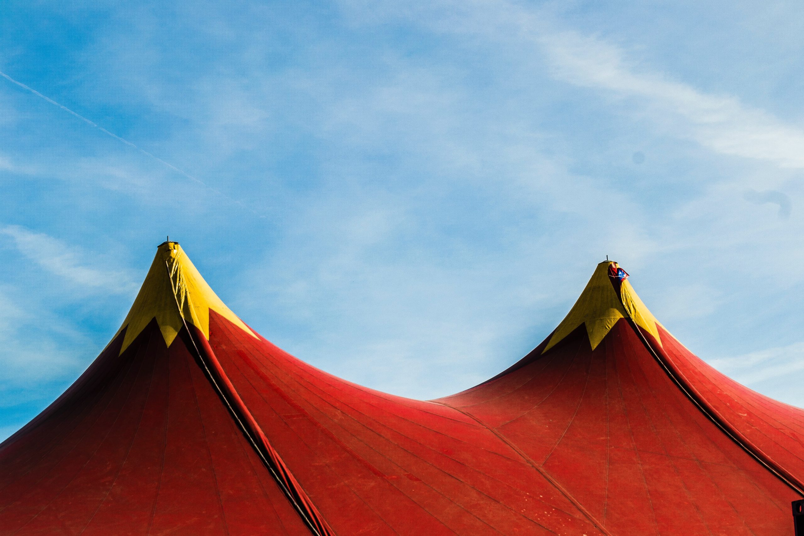 A photograph featuring two peaks at the top of a circus tent. The peaks are a bright scarlet colour and the tips are a mustard yellow. Above the tent, the sky is bright blue with a subtle dusting of white clouds.