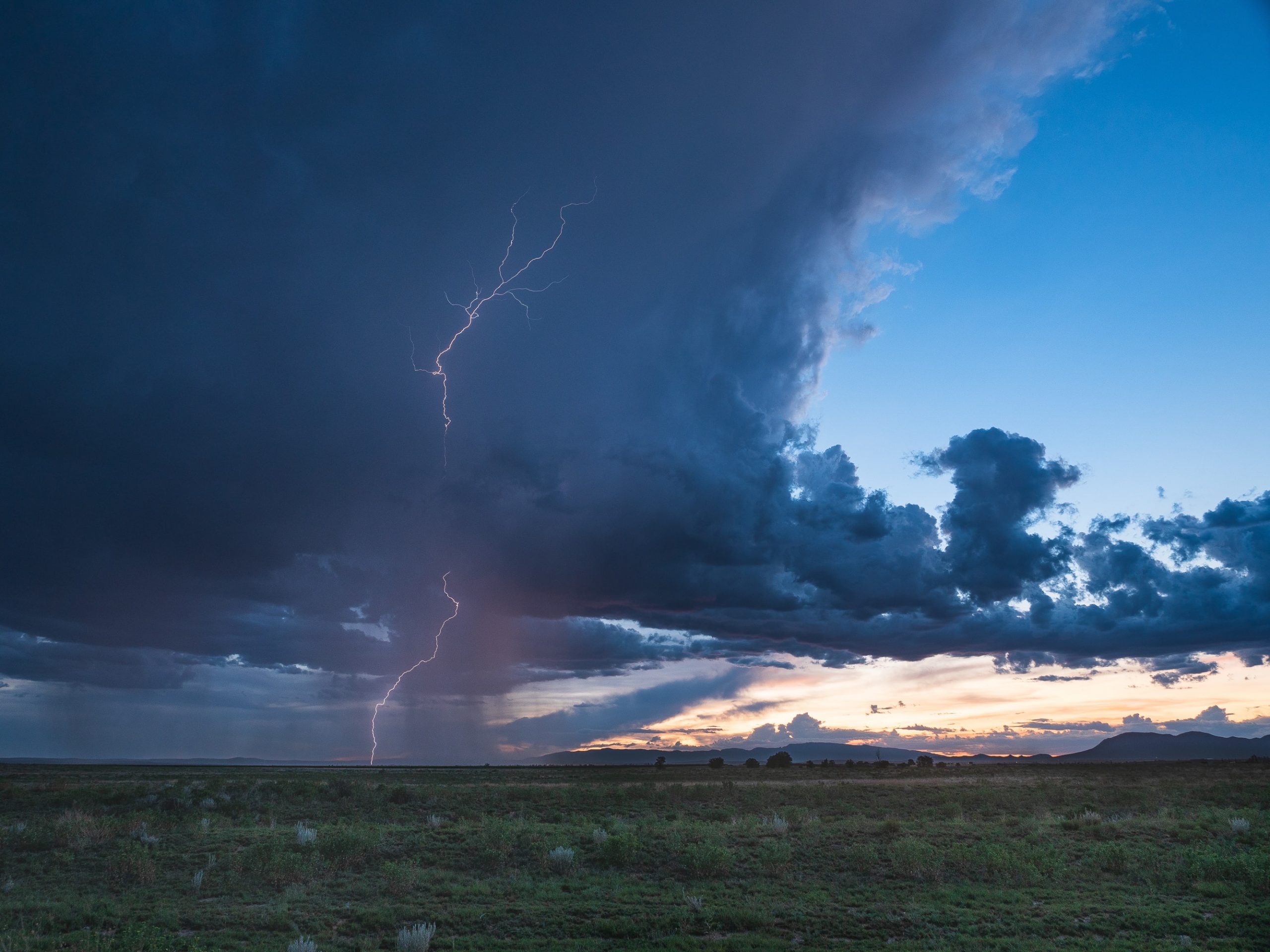 A photograph of a bolt of lightening striking an empty green field. The sky is filled with several dark clouds heavy with rain. In the right side of the photo, there is a gap in the clouds where a deep blue sky and a yellow-orange sunset are peeking through.