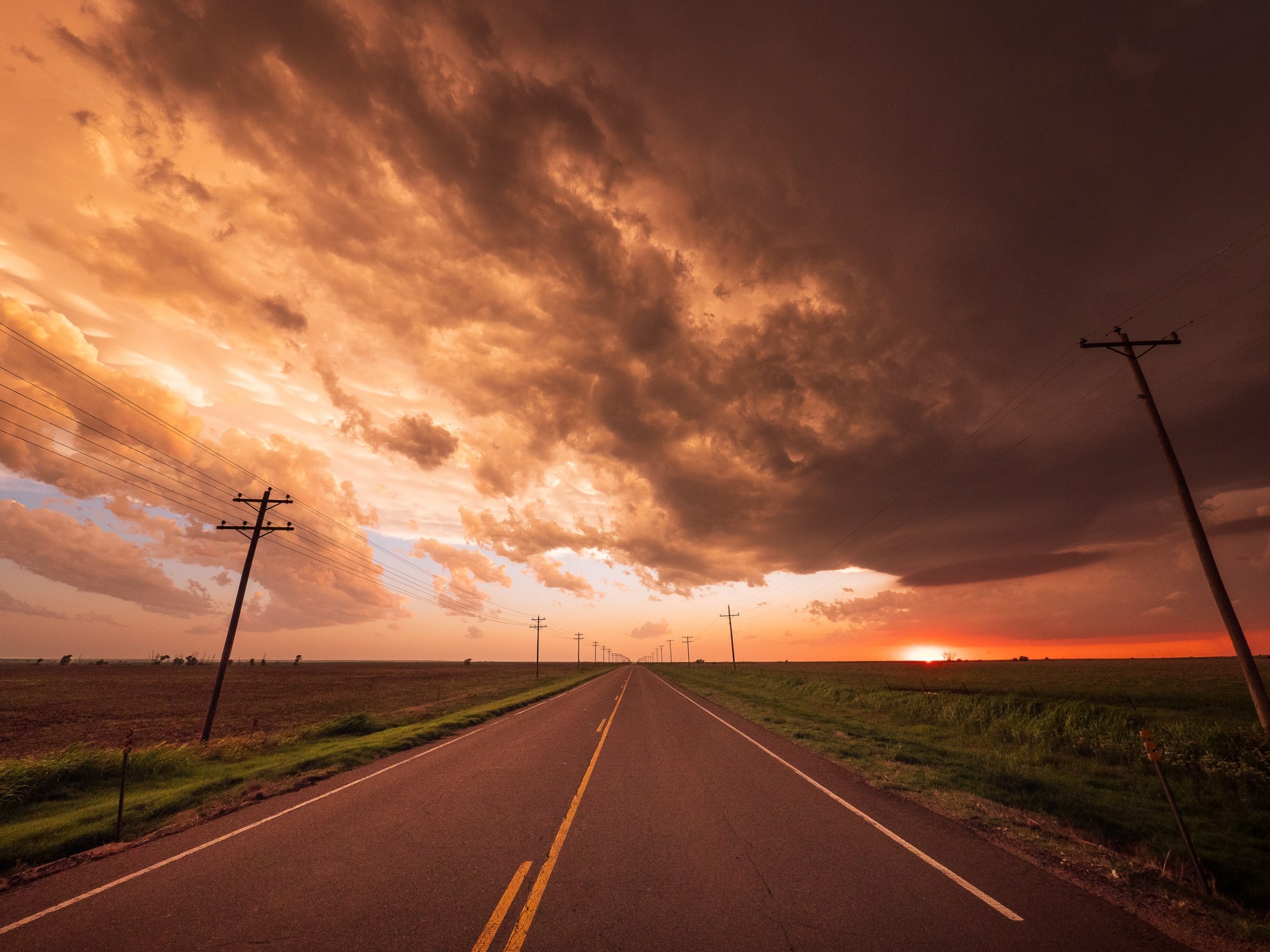 A photograph of a long, straight road lined with utility poles going off into the distance. On either side of the road are empty green fields. The sky is filled with dark grey clouds which are coloured by the golden-orange sunset. The sun is also depicted setting on the right side of the photo.