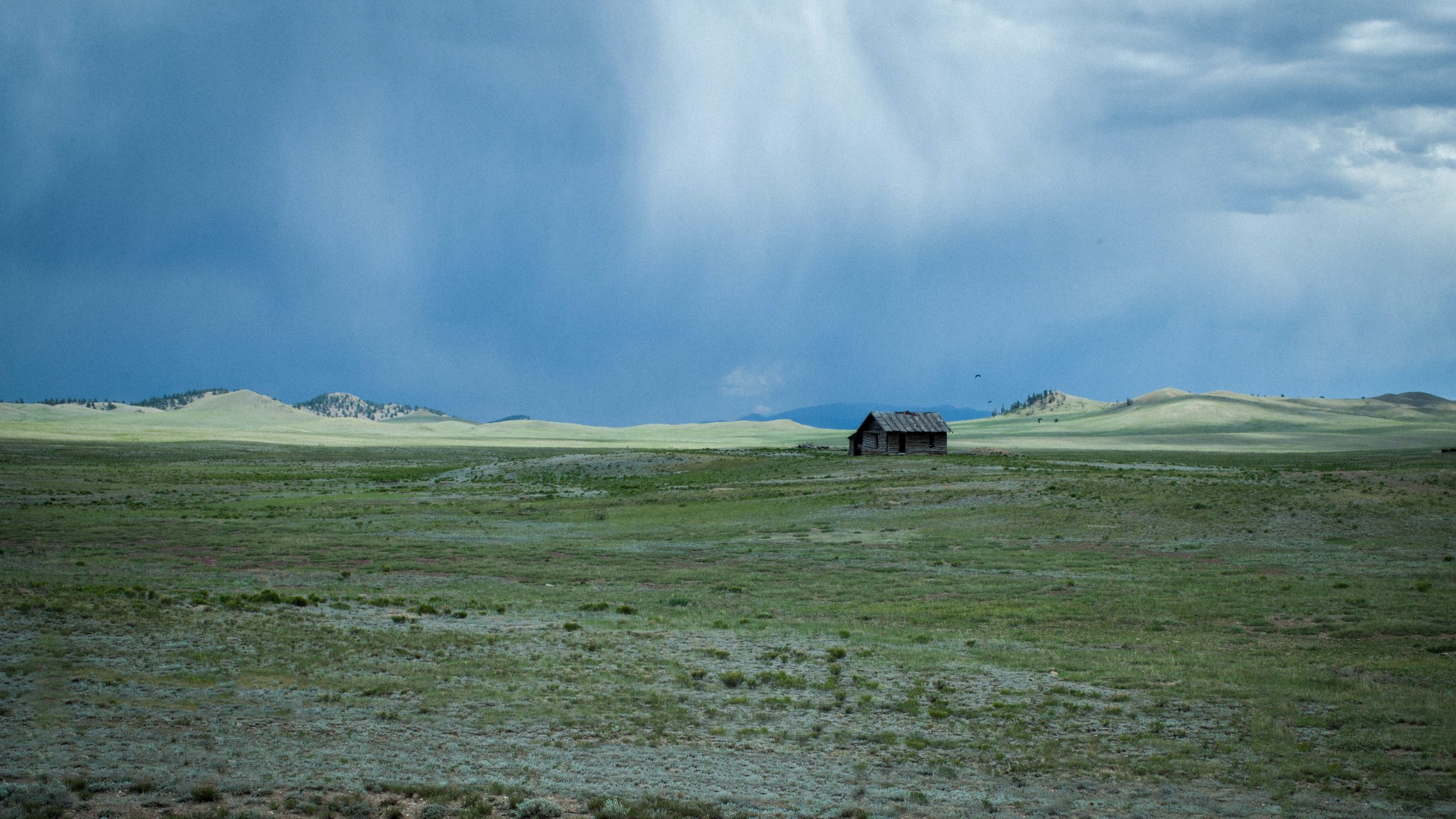 Photographie d'un grand champ vert entouré de collines en pente. Au centre du champ se trouve une petite cabane marron. Au-dessus, le ciel est d'un bleu froid et sombre.