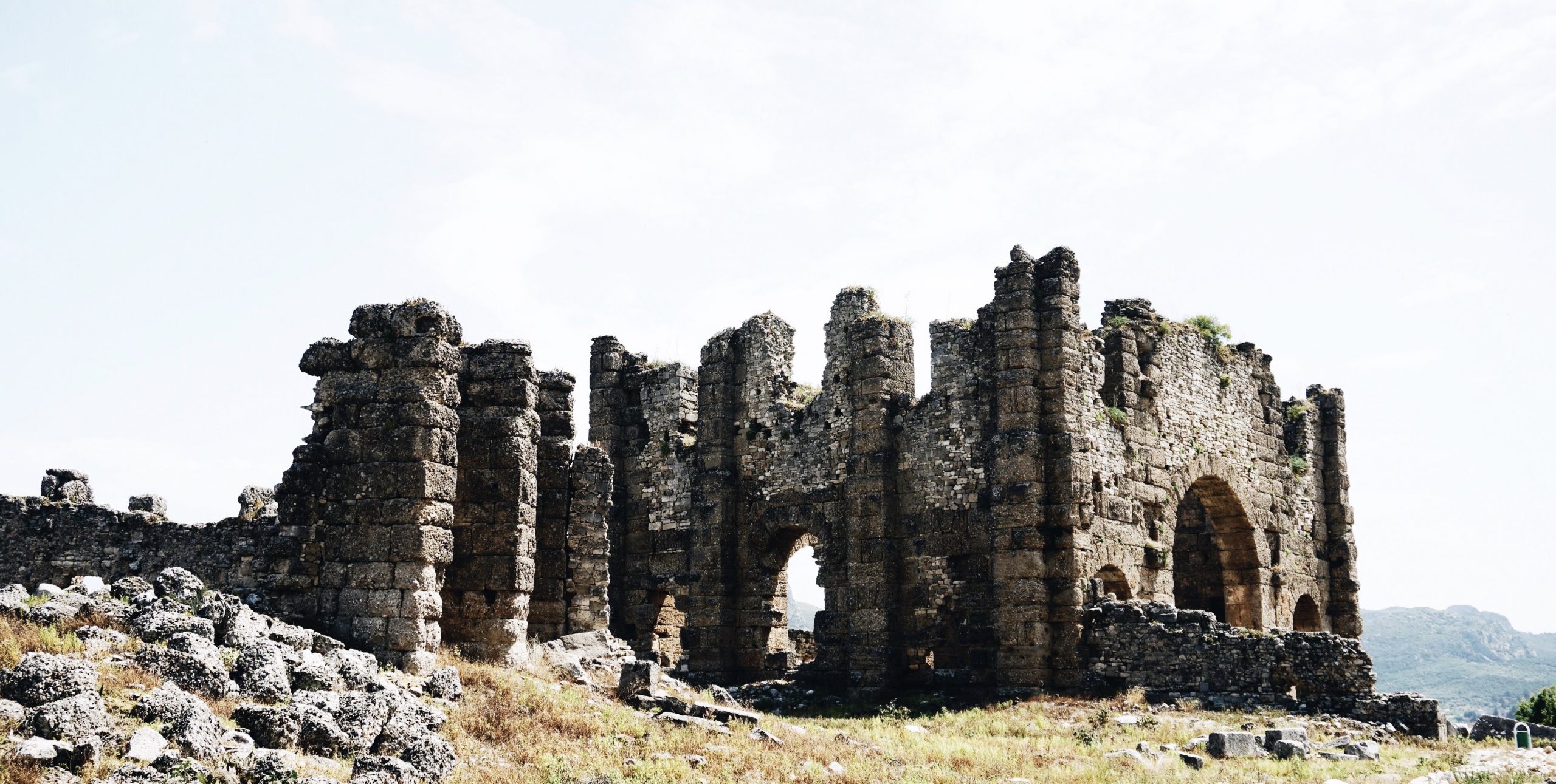 Photographie d'une ruine de château en ruine reposant au sommet d'une colline verte et herbeuse. Les restes de quatre murs en pierre, ainsi que deux portes rondes sont visibles sur le côté droit et à l'arrière de la ruine. Autour de la ruine se trouvent des amas de pierres et de rochers gris. Le ciel calme est d'un bleu pâle.