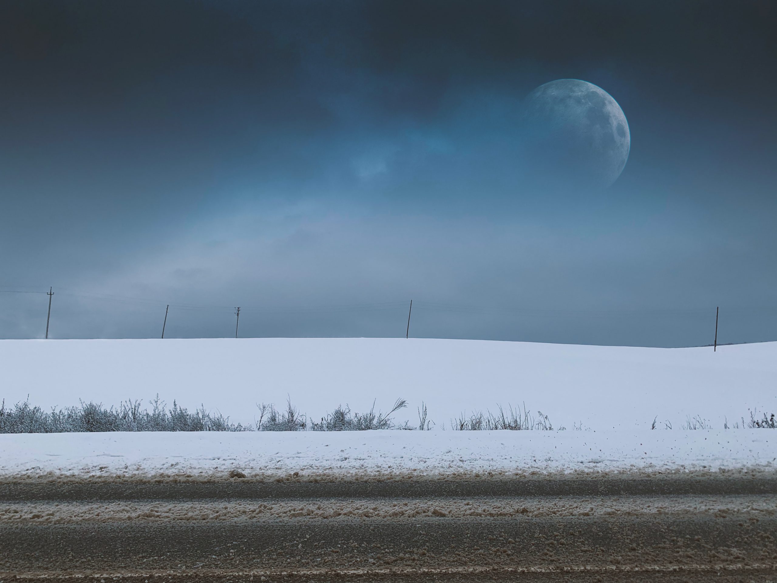 A photograph of a snow-covered field with tufts of grass peeking through. Above, the sky is a dark blue with smoky wisps of clouds. To the right, a full moon peers through the clouds.