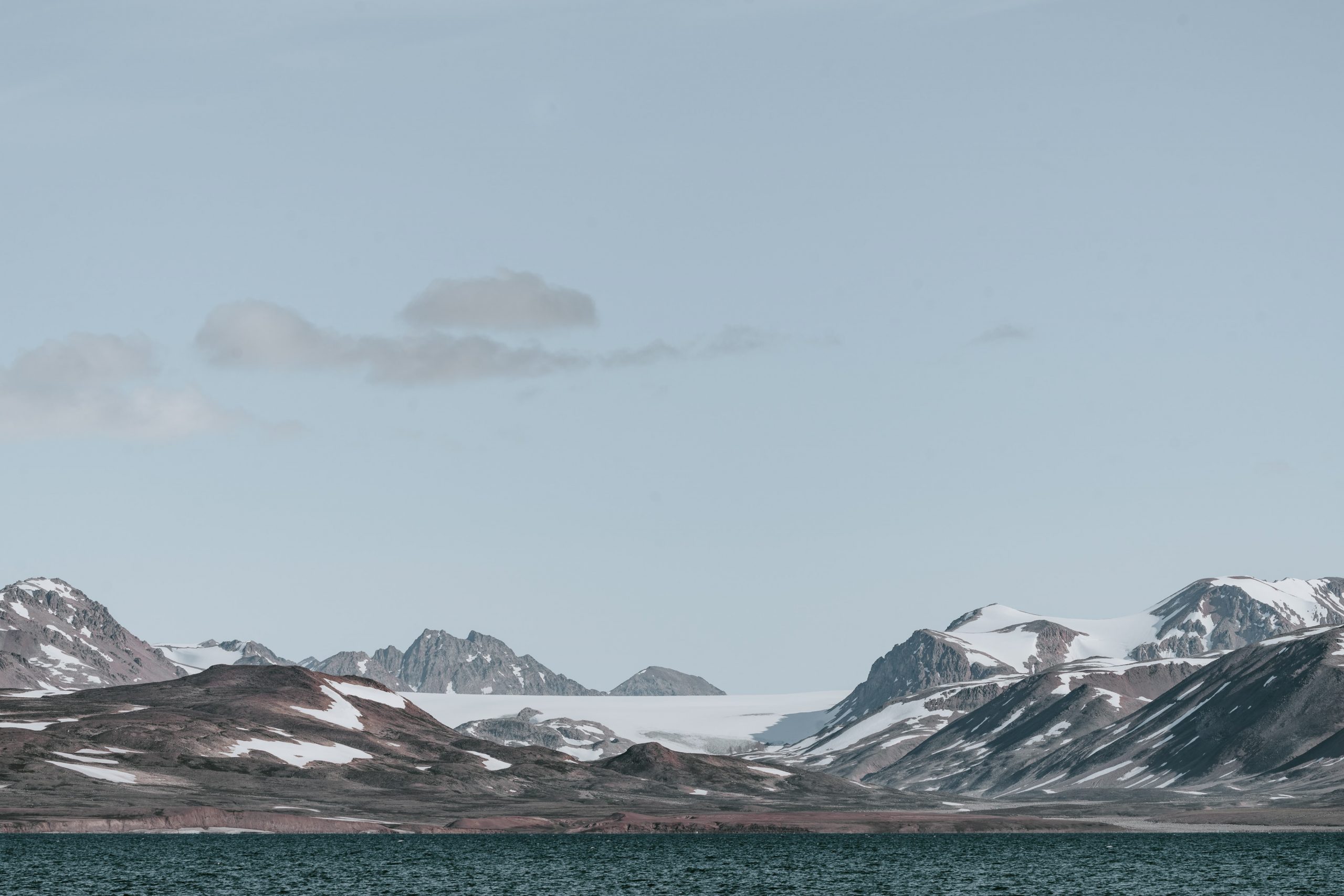 A photograph of a dark blue ocean with jagged, snow-dusted mountains in the background. The sky is a frosty blue with wisps of grey clouds in it.