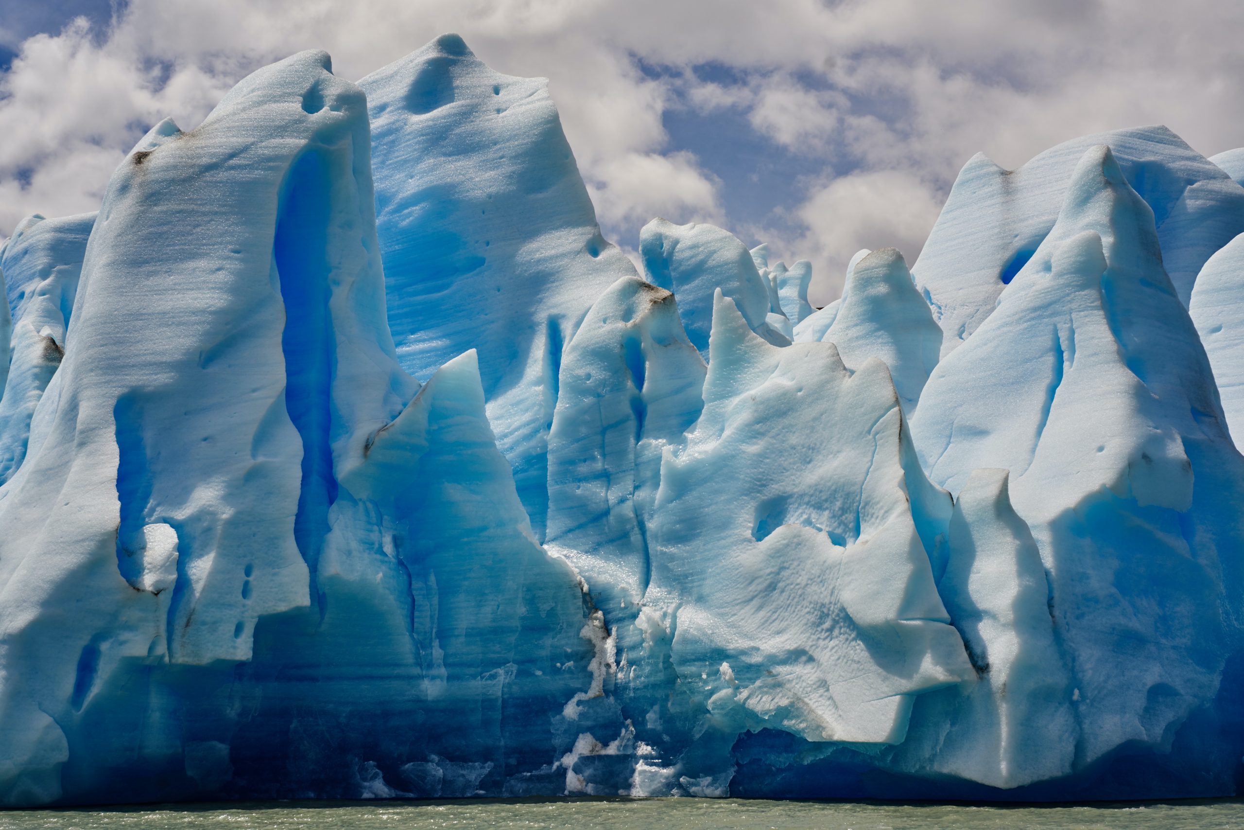 A close-up photograph of tall, white-blue icebergs. The sky above is a happy light blue with puffs of fluffy white clouds.
