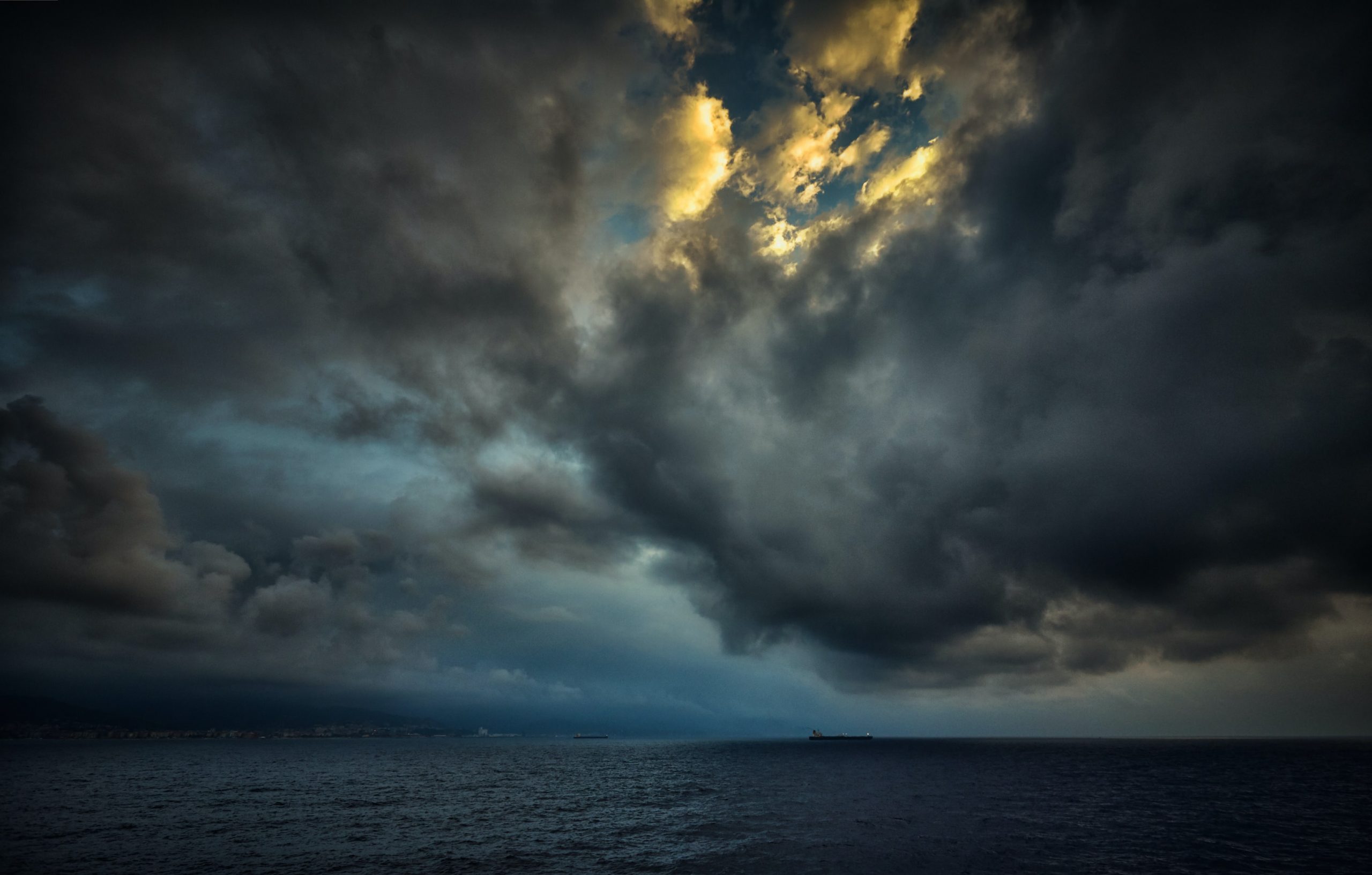 A photograph of a midnight black, calm ocean. Above, the sky is a dark blue and is filled with smoky grey storm clouds. In the centre of the cloud cluster, a hopeful spot of sunshine peeks through.