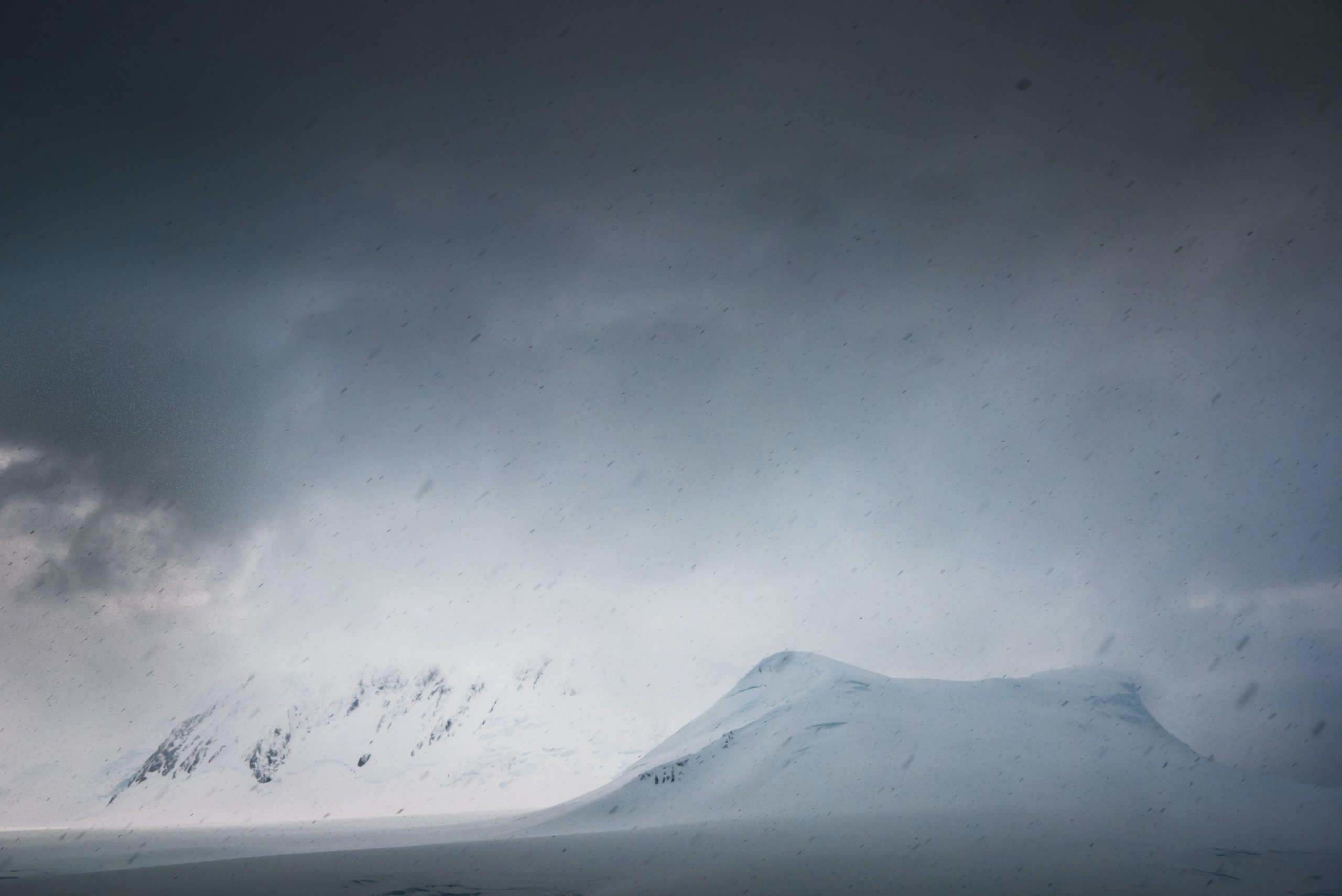 Une photographie de montagnes rigides couvertes de neige. Le ciel est rempli de nuages de neige sombres, et un vent fort souffle de la neige dans l'air froid.